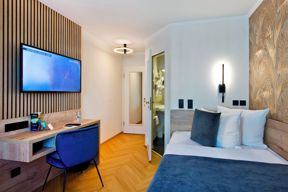 Una habitación de hotel moderna con un escritorio de madera, un televisor de pantalla plana, una cama con cabecera y una lámpara fijada a la pared.