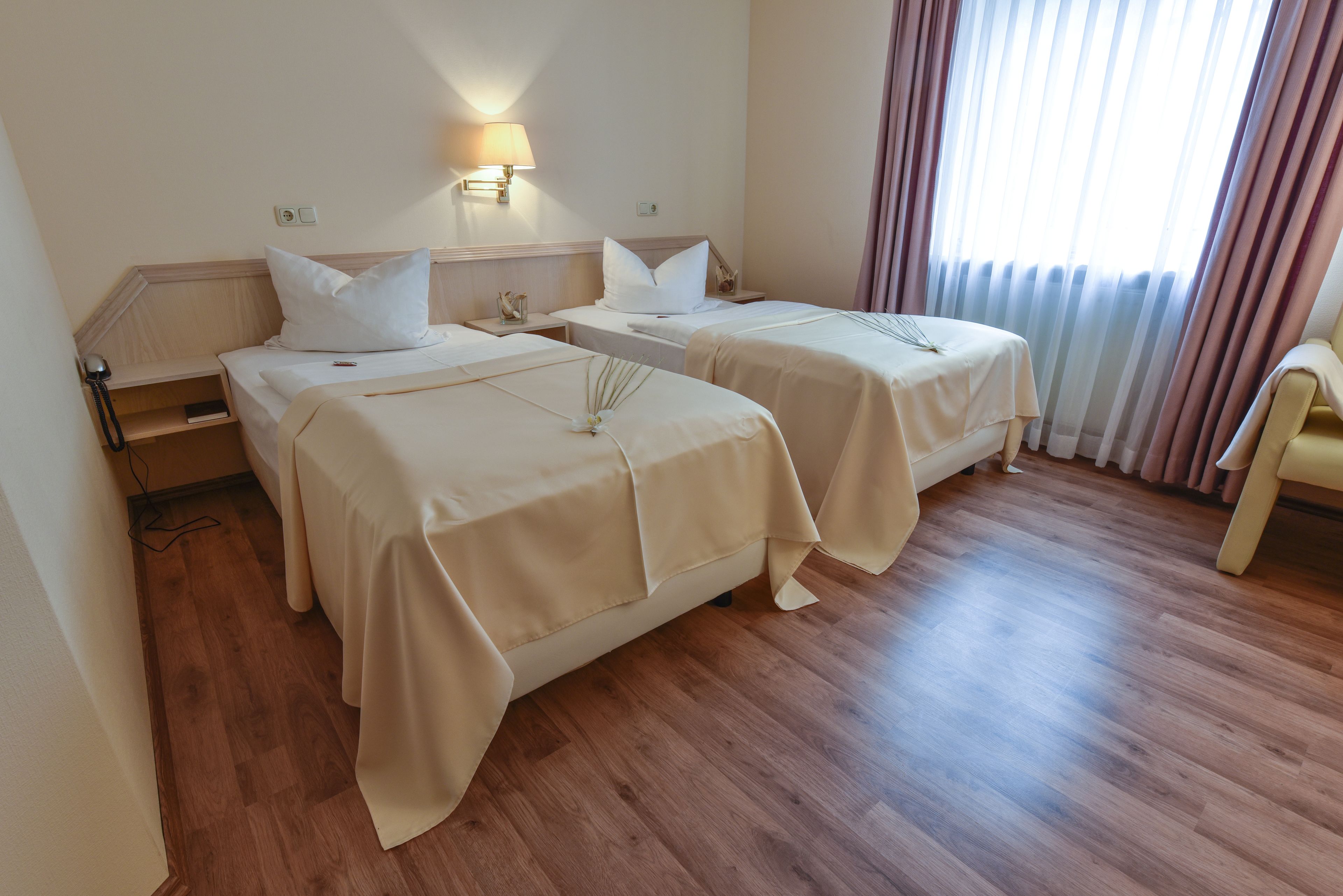 Habitación de hotel con dos camas, suelo de madera, una mesita de noche con lámpara y una ventana con cortinas.