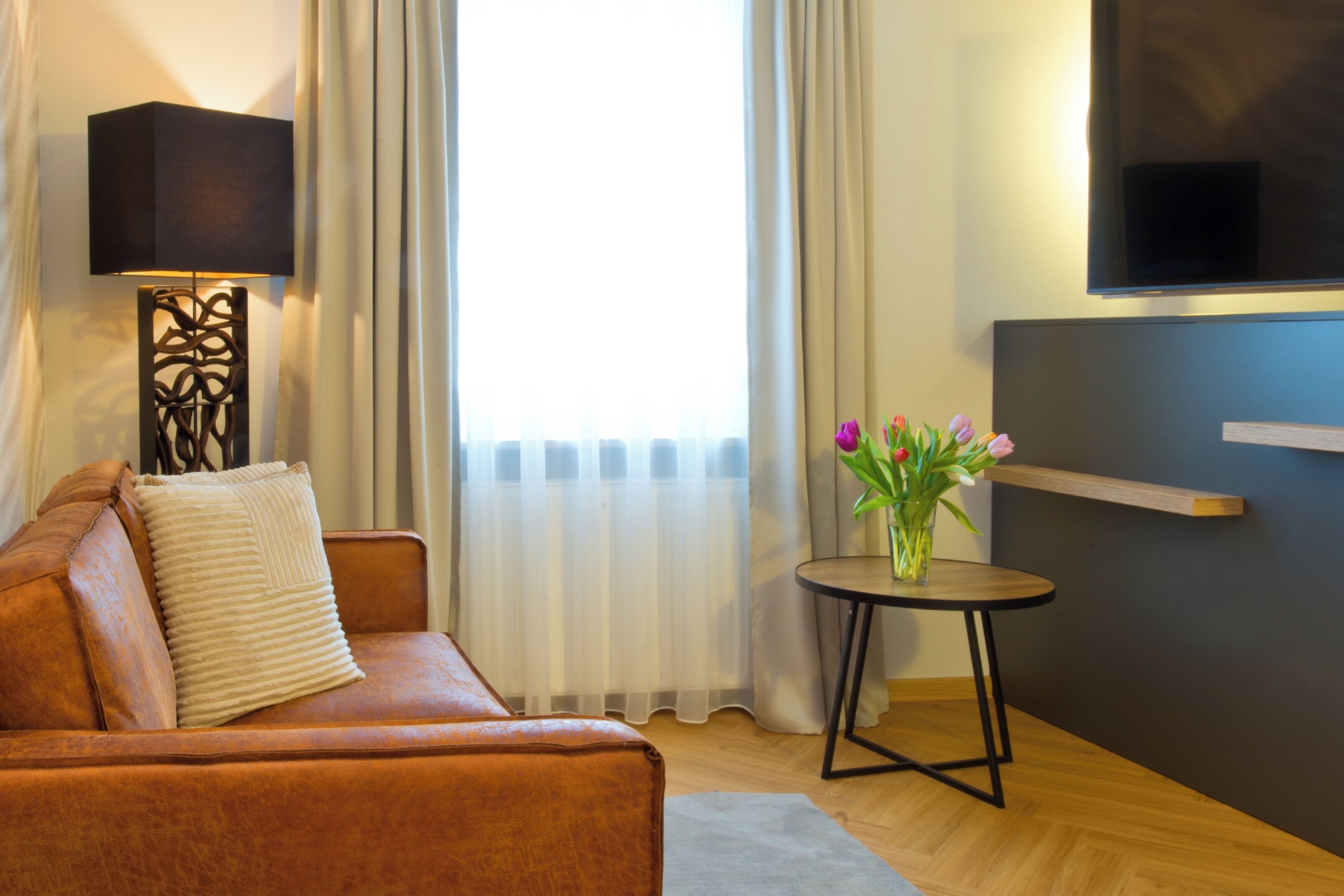 Una habitación de hotel moderna con un sofá de cuero, una mesita redonda con un florero, una lámpara de pie y un televisor de pantalla plana montado en la pared.
