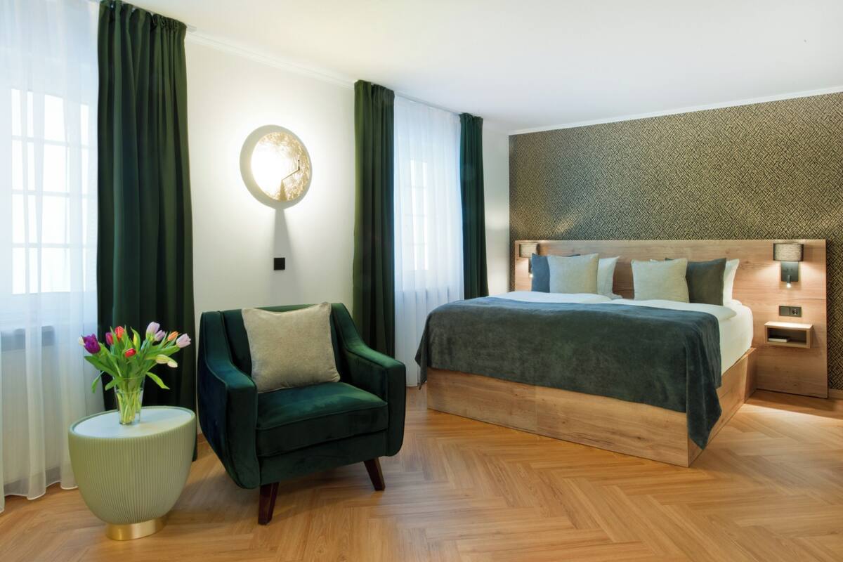 Un dormitorio moderno con una cama grande, un sillón verde, una mesita redonda con un jarrón de flores y una lámpara de pared.