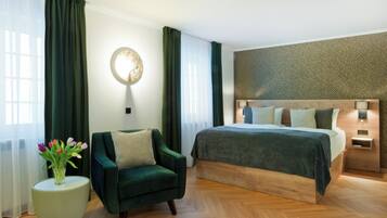 A modern bedroom with a large bed, a green armchair, a round side table with a vase of flowers, and a wall-mounted light fixture.