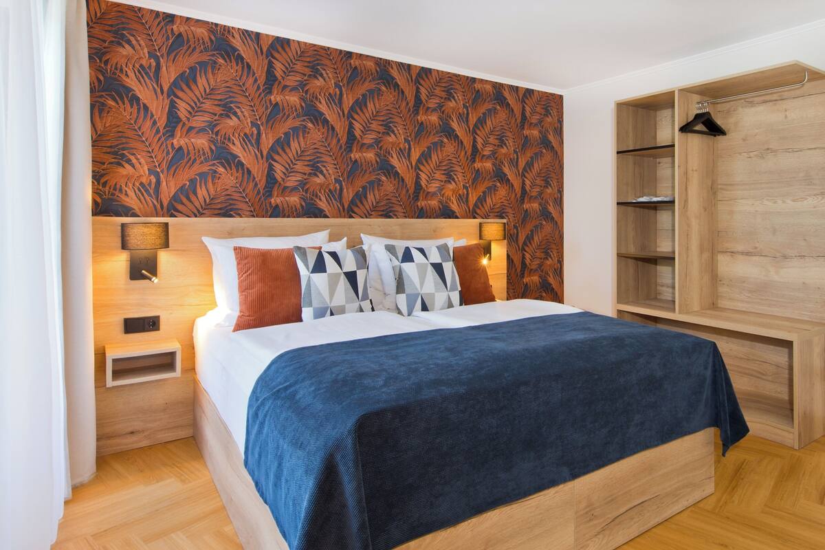 Una cama cuidadosamente tendida con una colcha azul, un cabecero de madera y almohadas estampadas. Hay una mesita de noche con una lámpara y un armario de madera con estantes.