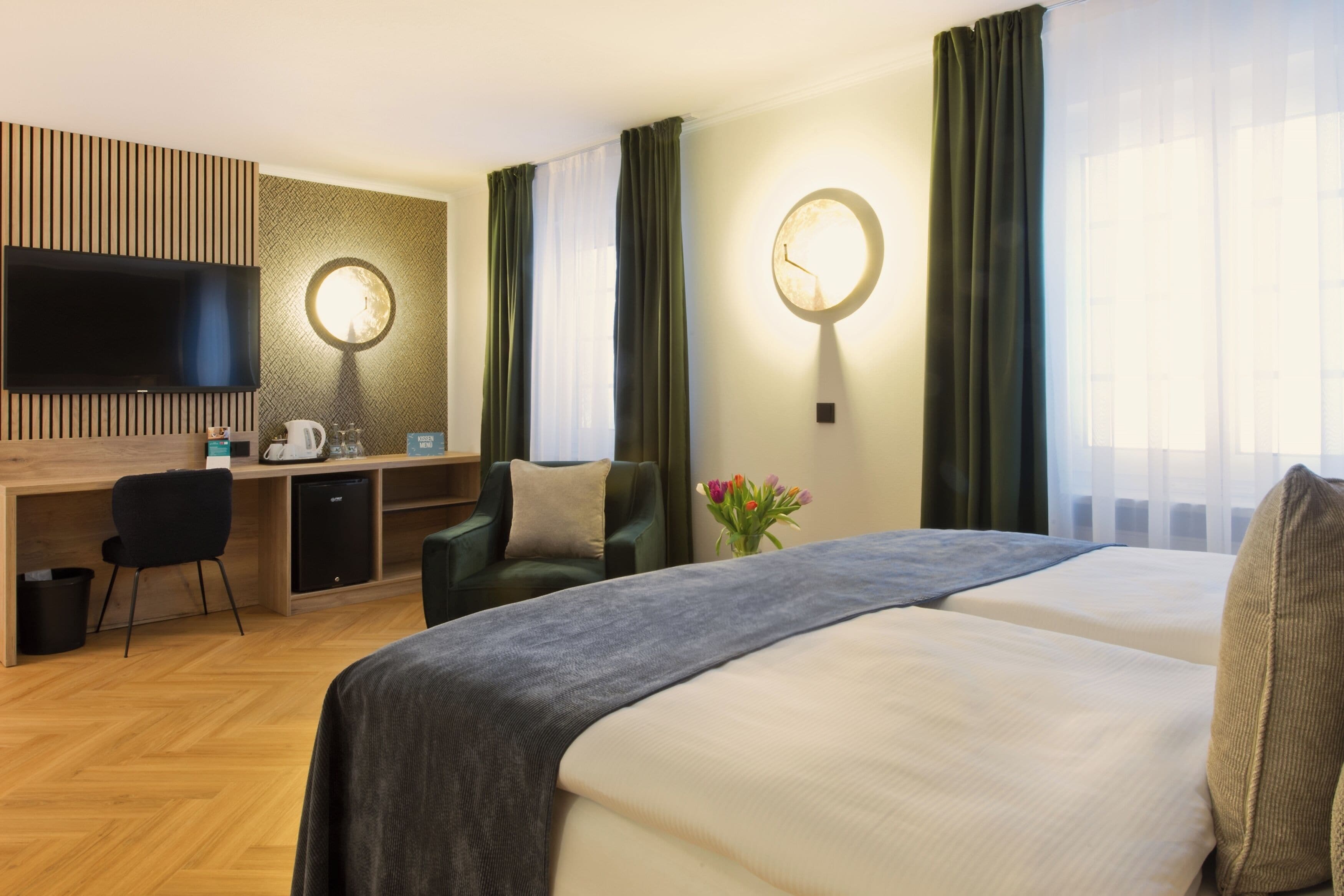 A hotel room with a bed, a desk with a chair, a television, and a clock on the wall.