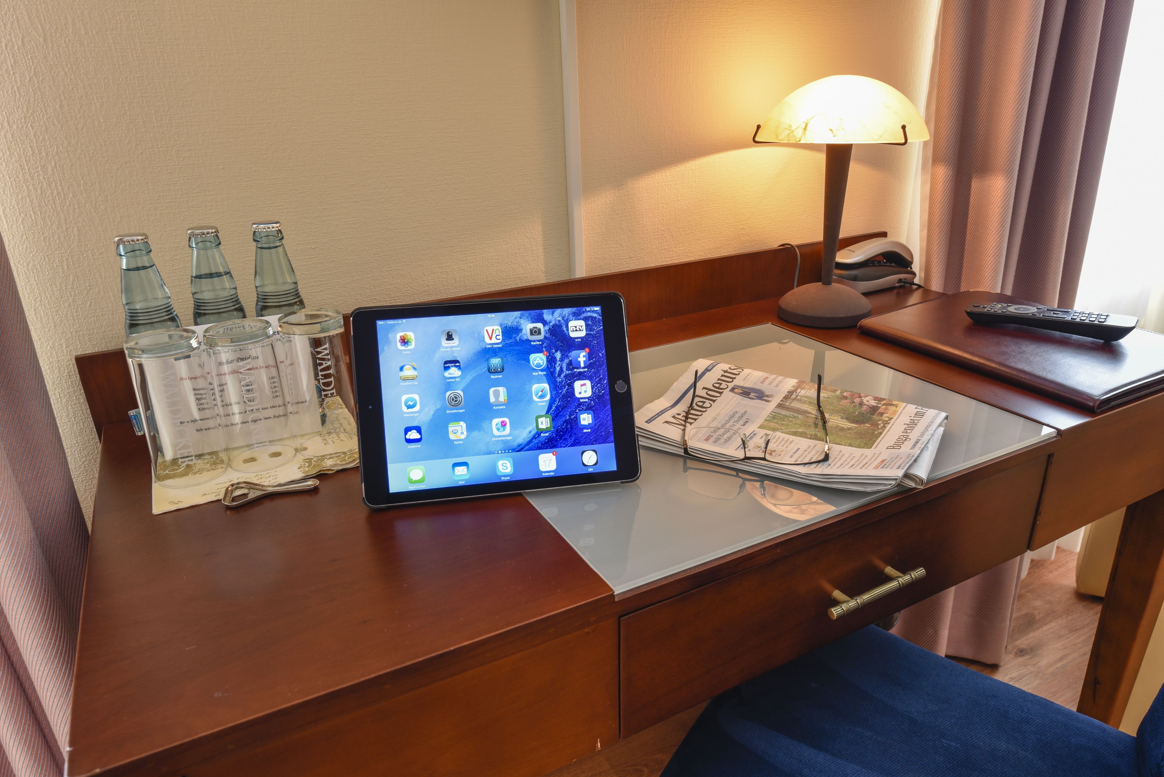 Et hotelbord med en tablet, flasker, en lampe, en fjernbetjening og en avis.