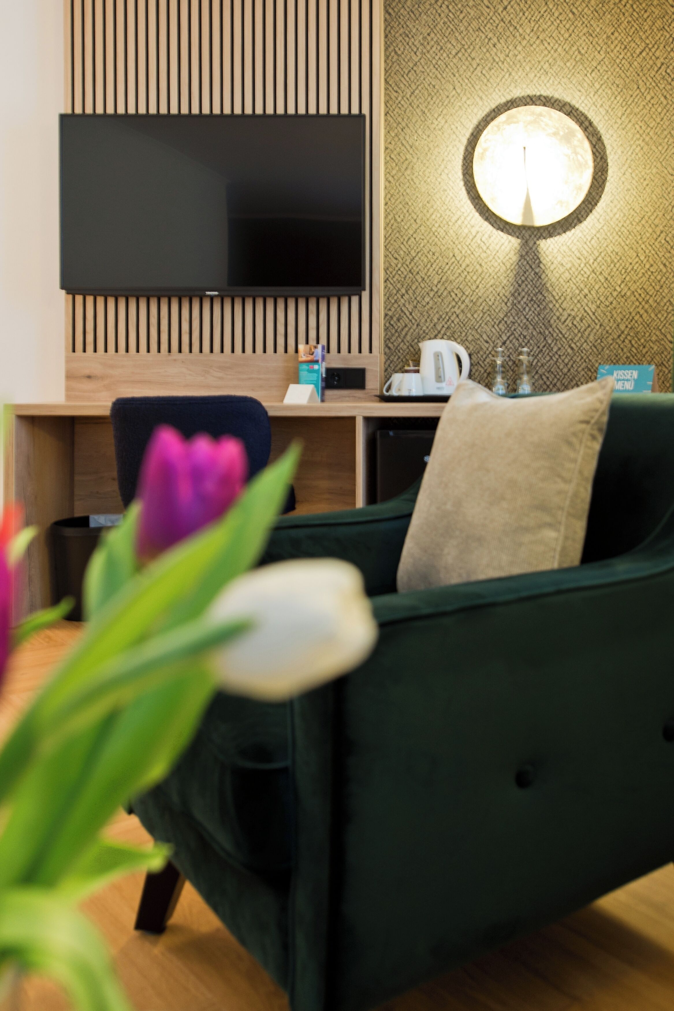 Ein modernes Zimmer mit einem Flachbildfernseher an einer Wand aus Holzpaneelen, einem runden Tisch mit einer Lampe, einem grünen Sofa mit Kissen und einem kleinen Couchtisch.