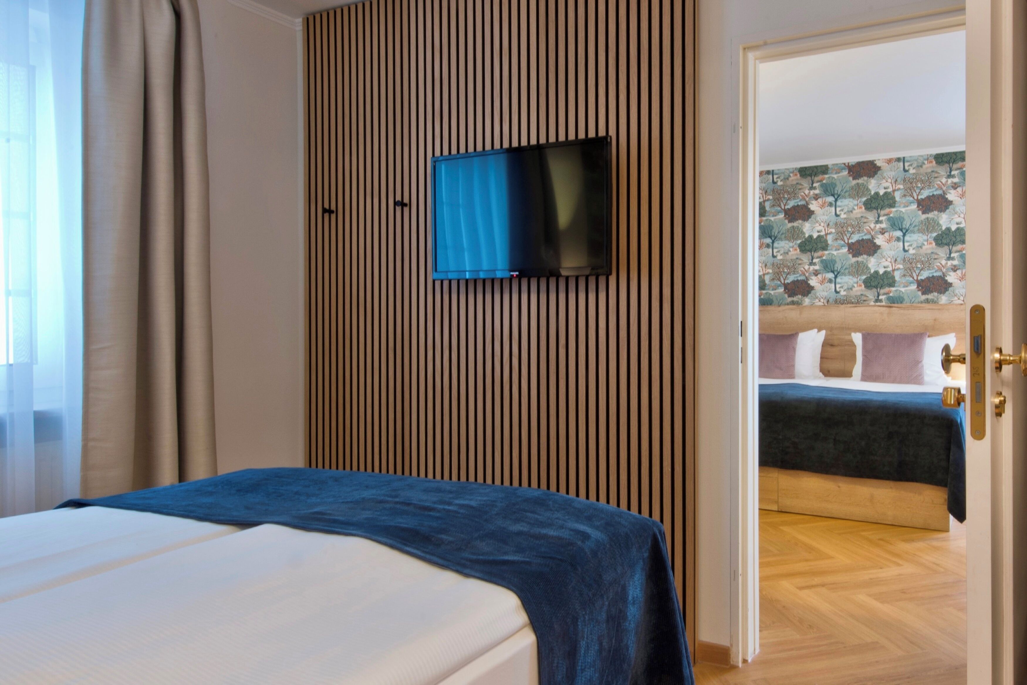 Habitación de hotel con una cama, un televisor instalado en una pared de paneles de madera y una puerta que conduce a otra habitación.