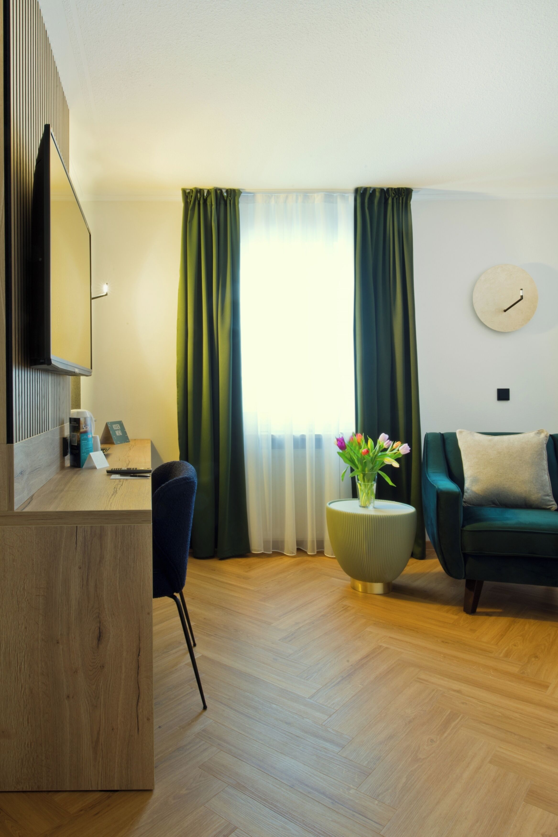 Una habitación de hotel moderna con un escritorio de madera, una silla cómoda, un sofá verde y una mesa redonda con un jarrón de flores.