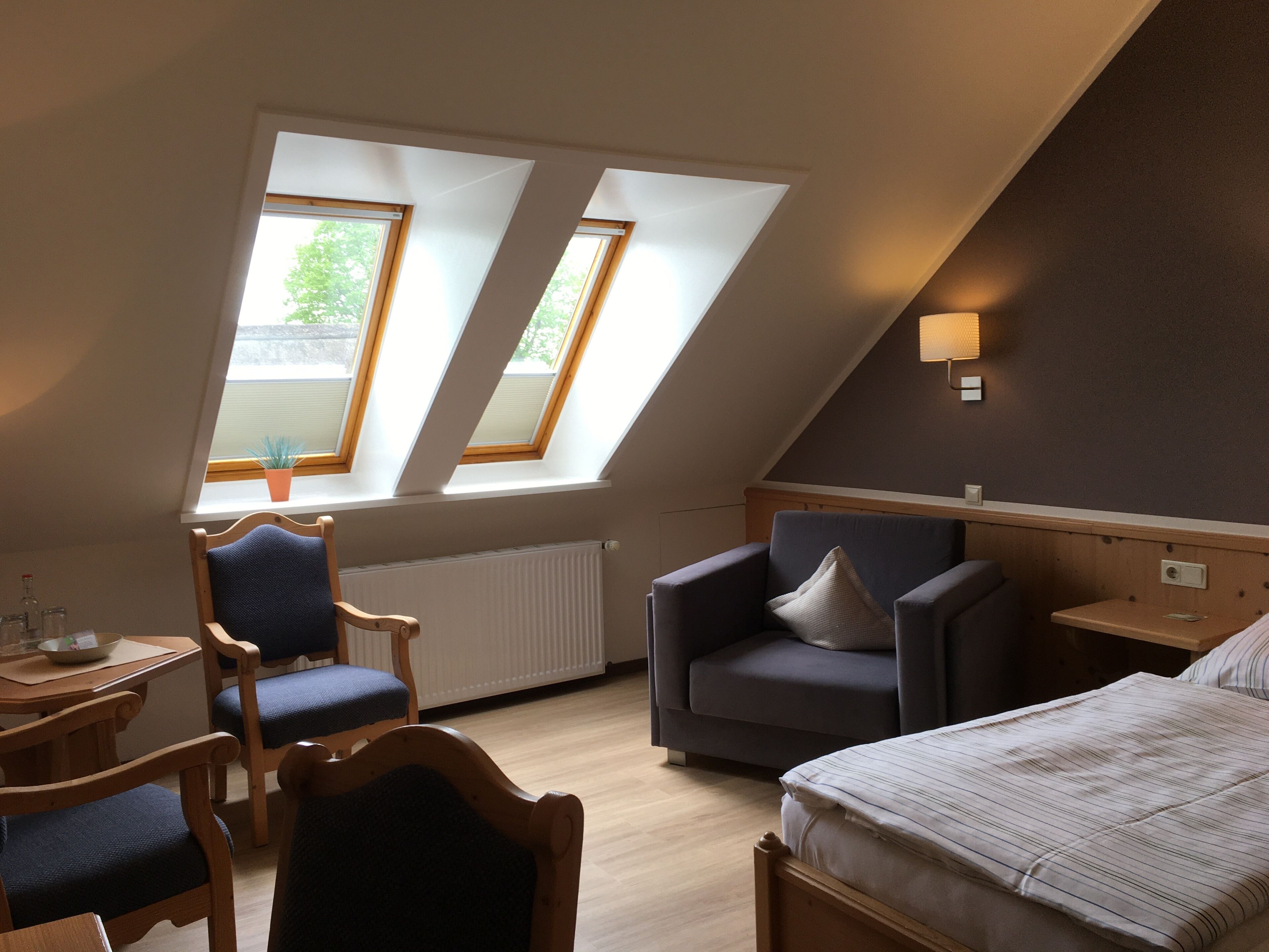 A room with a sloped ceiling, two skylights, a bed, a chair, a table, and a radiator.