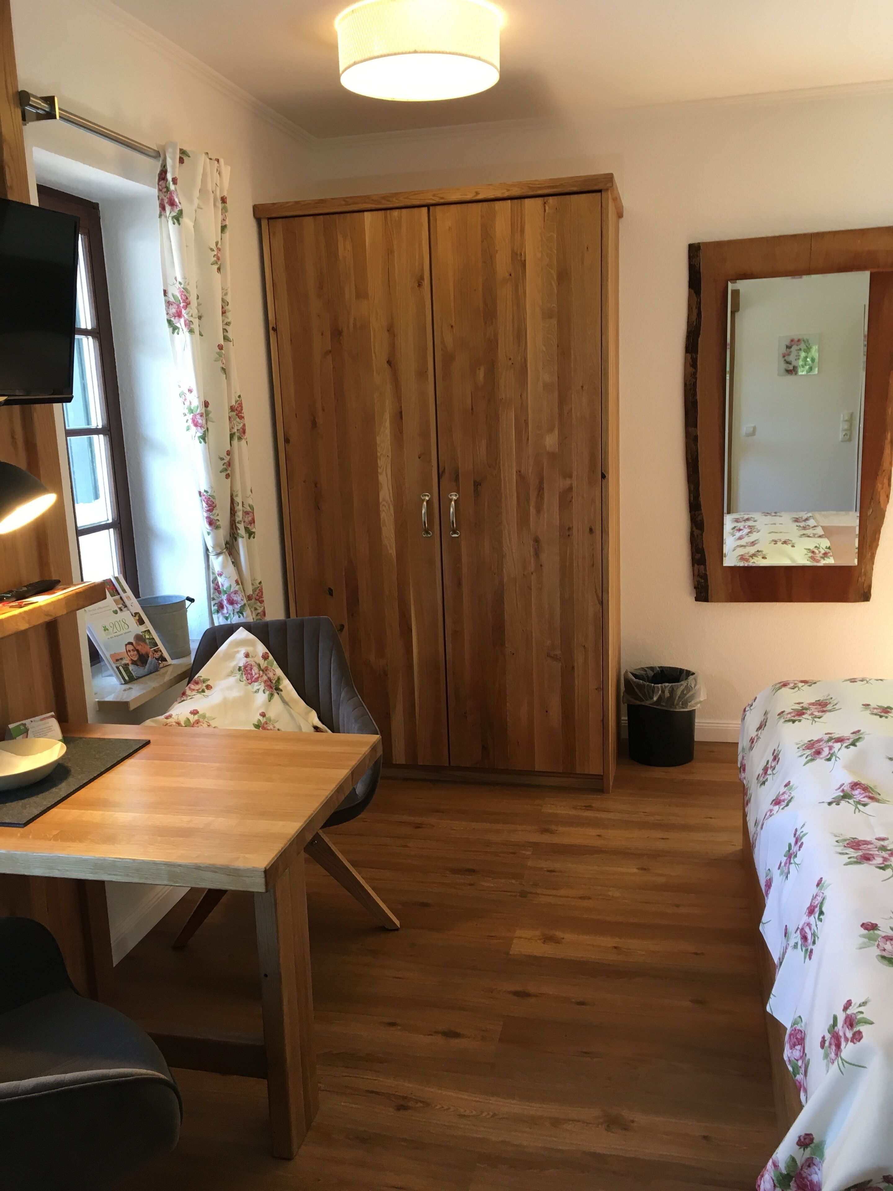 Ein Zimmer mit Holzschrank, einem Bett mit Blumenmuster-Bettwäsche, einem Tisch mit Stuhl, einem Fernseher und einem Fenster mit floralen Vorhängen.