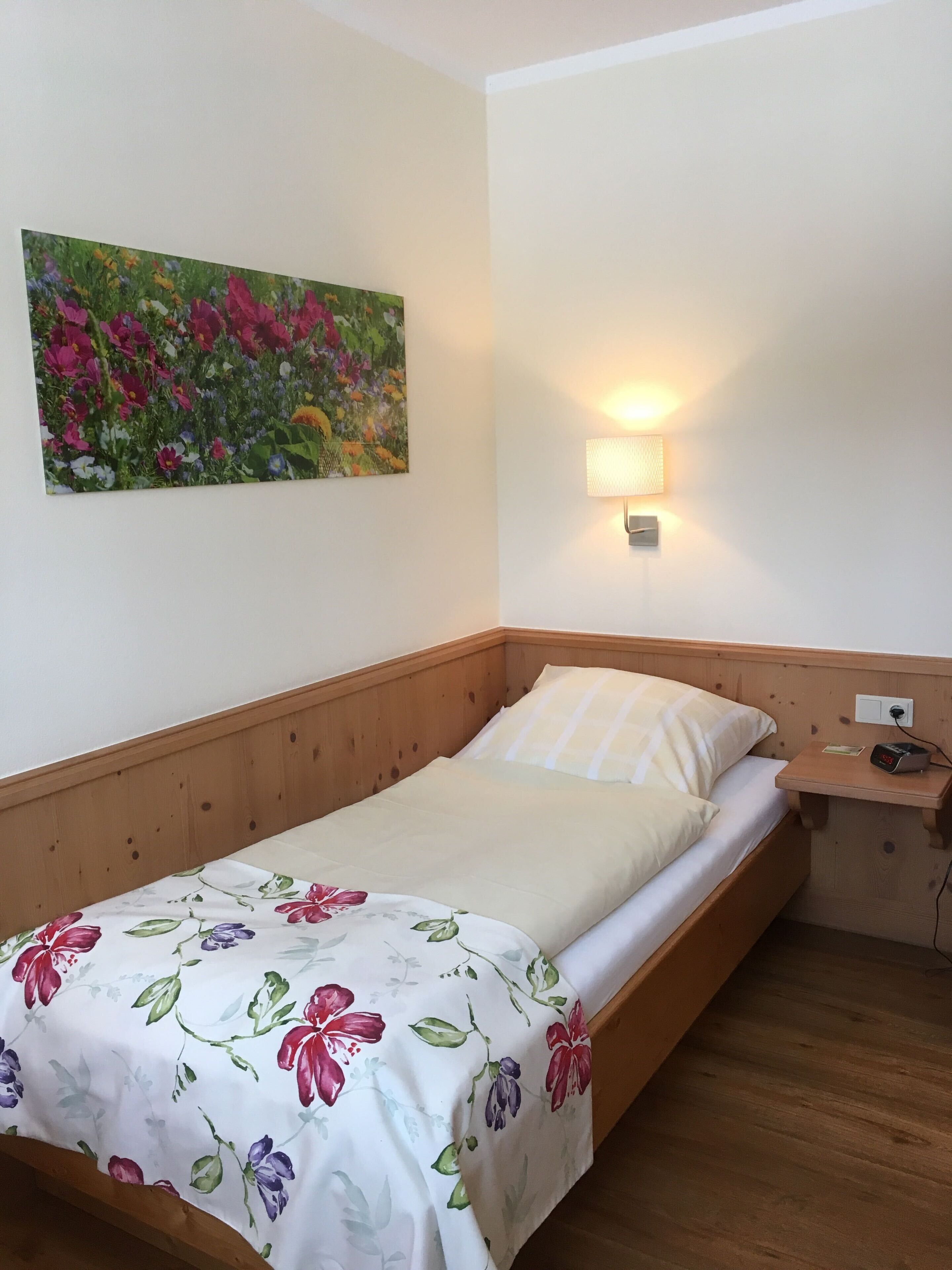 Ein Einzelbett mit floralem Überwurf, ein Nachttisch mit Lampe und ein Blumenbild an der Wand.