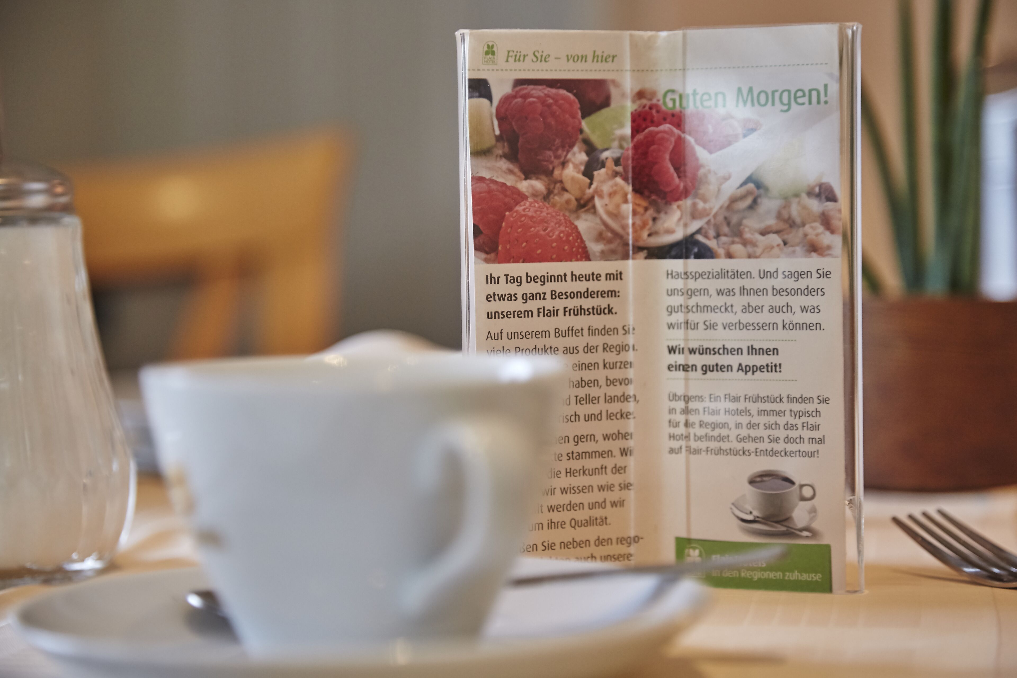 Eine Speisekarte mit Text und Abbildungen von Speisen, platziert auf einem Tisch mit Tasse und Untertasse.
