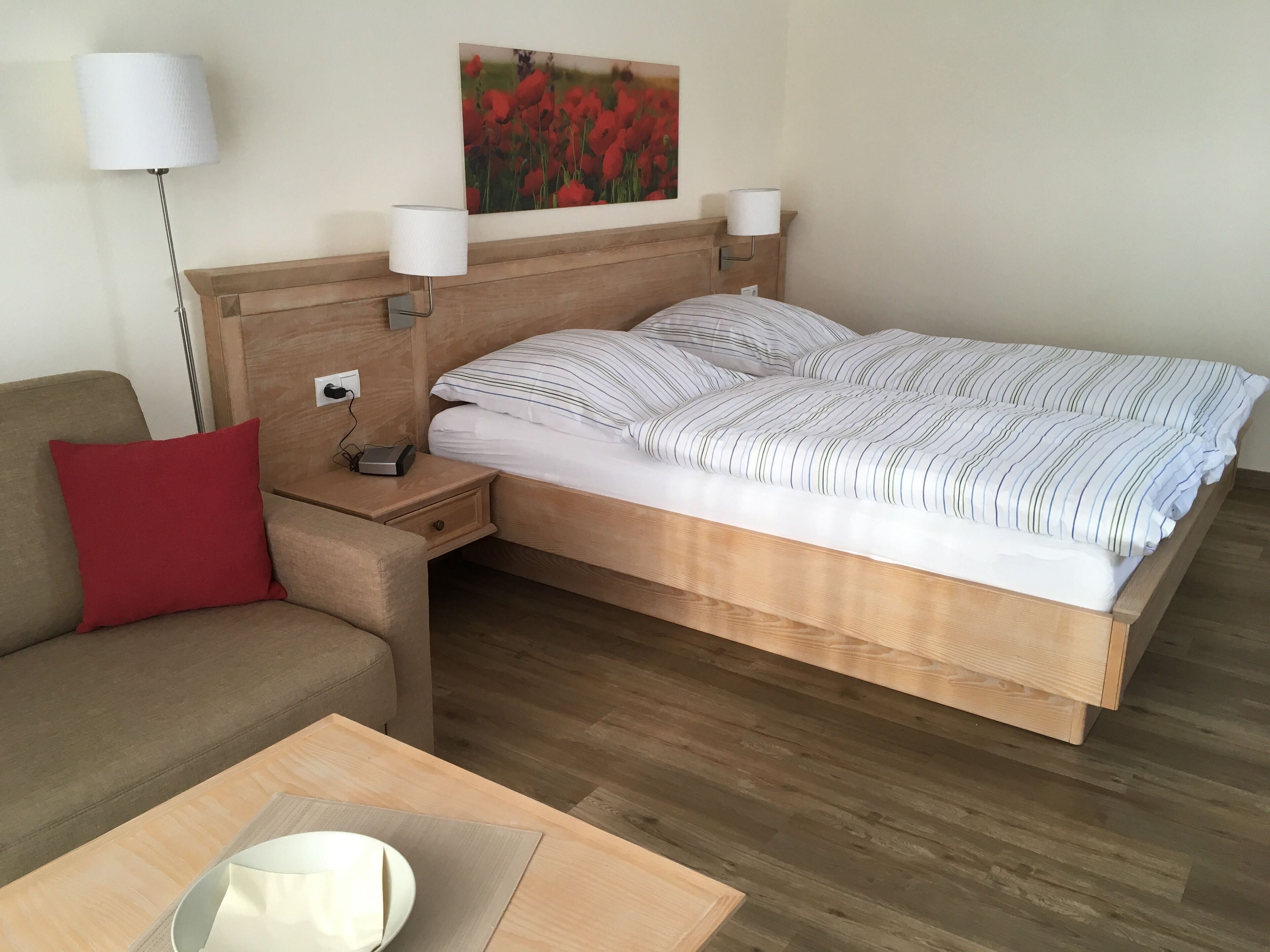 A bedroom with a wooden headboard, a bed with striped bedding, a red pillow on a sofa, a bedside table with a lamp, and a painting of red flowers on the wall.