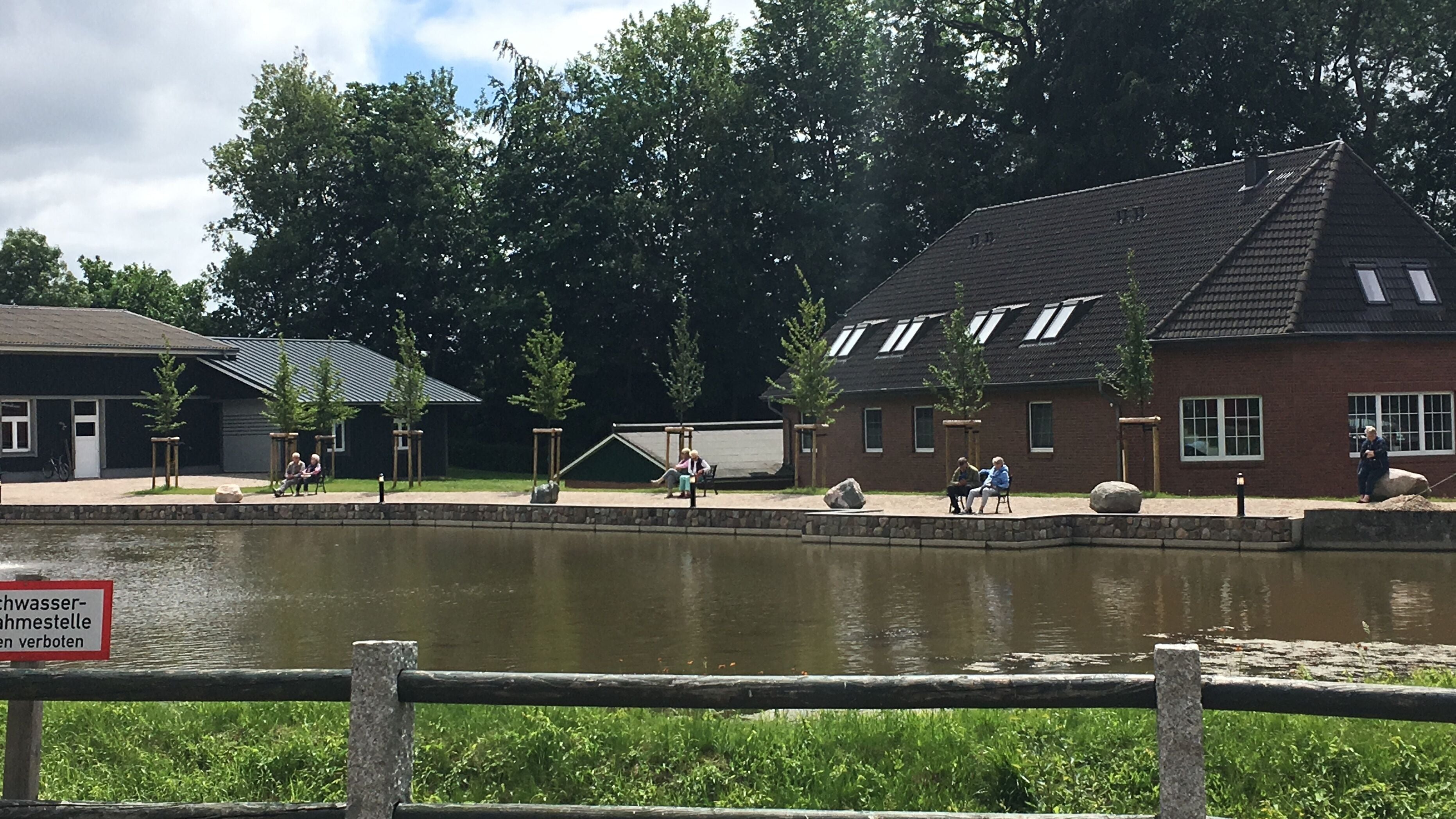 Un área junto al río con un edificio de ladrillo, una valla de madera y gente sentada junto al agua.