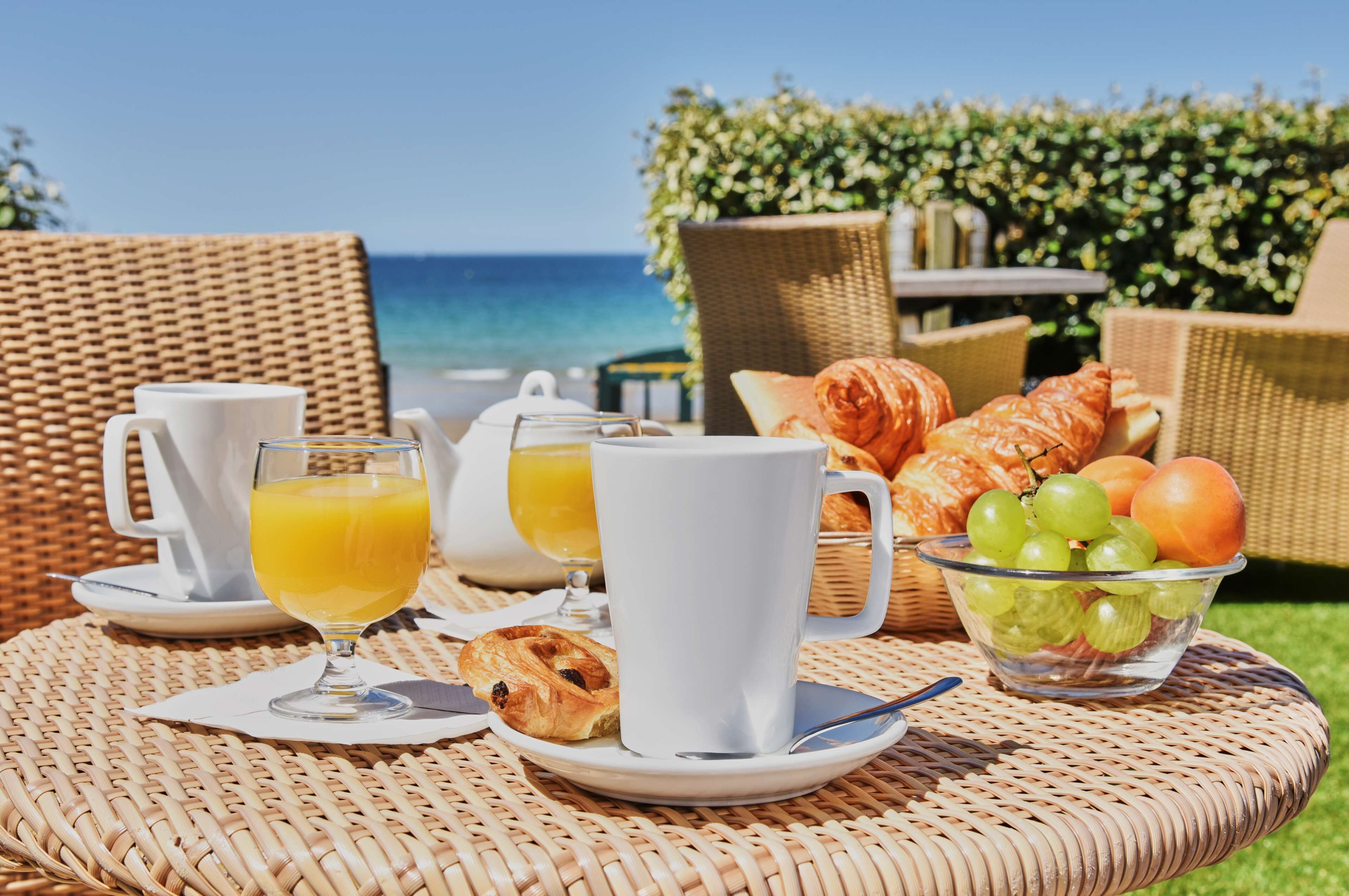 A breakfast setup with croissants, a bowl of grapes, and orange juice on a wicker table with a view of the ocean.