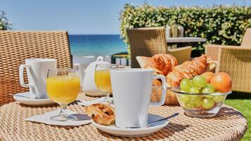 A breakfast setup with croissants, a bowl of grapes, and orange juice on a wicker table with a view of the ocean.
