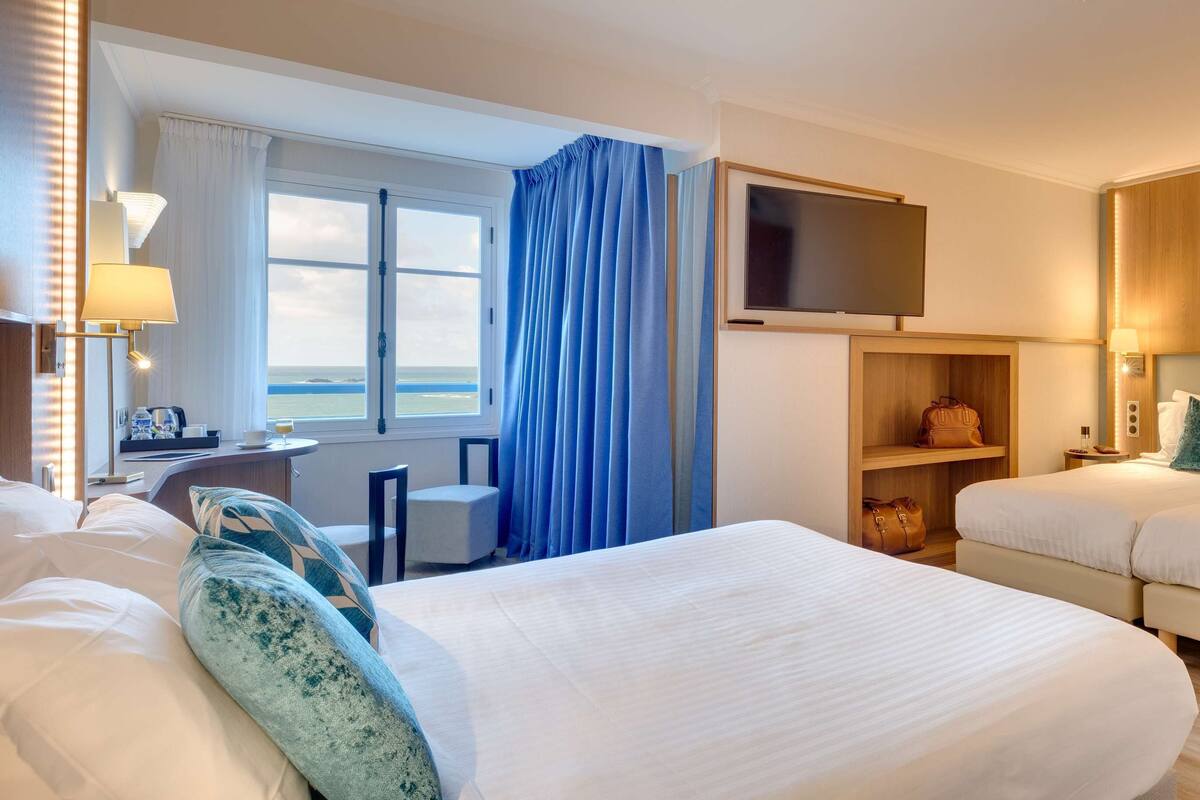 A hotel room with two beds, a desk, a TV, and a view of the sea.