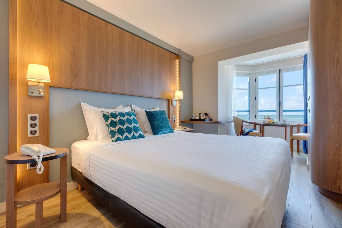 A hotel room with a large bed, bedside lamps, a small table with a phone, and a view of the sea through large windows.