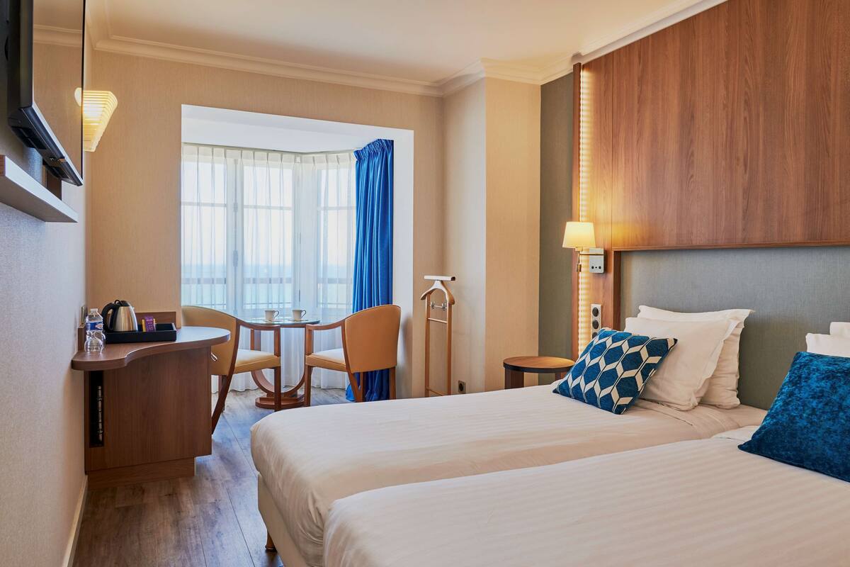 A hotel room with two beds, a desk with a chair, a small table, and a window with curtains.