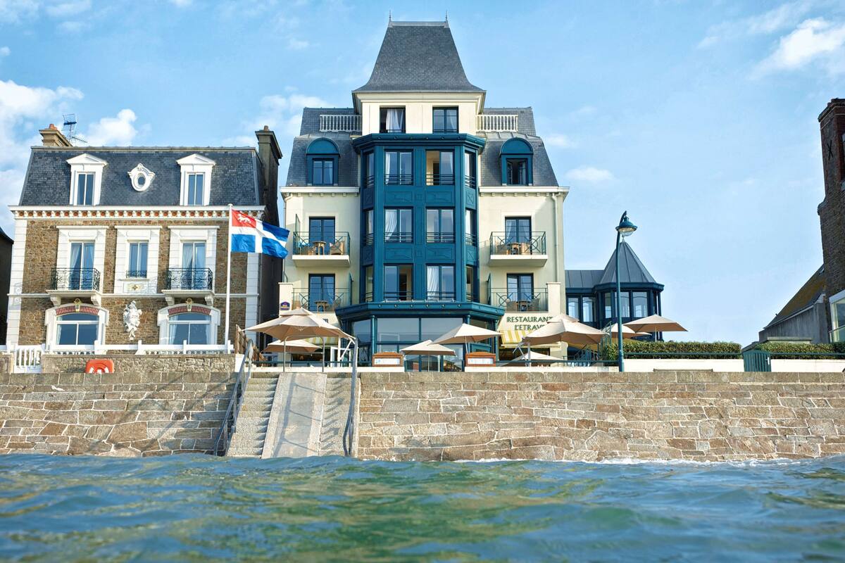 A waterfront hotel with a prominent corner building, outdoor seating, and a stone wall.