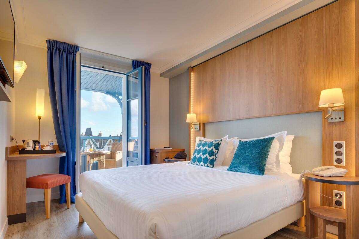 A hotel room with a large bed, a desk with a chair, a bedside table with a lamp, and a view of the outside.