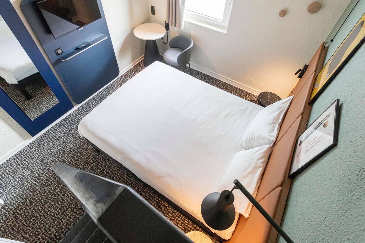 A hotel room with a bed, a desk, a chair, a small table, a lamp, a window, and a wall with a picture frame.