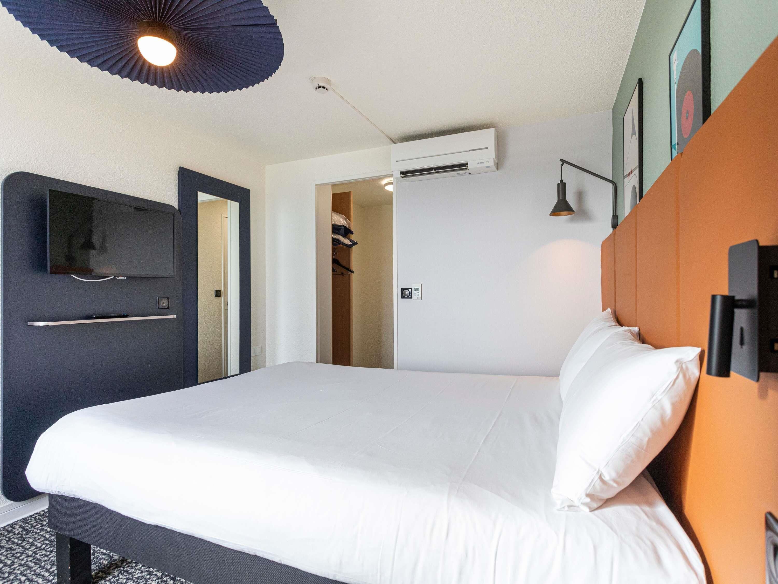 A hotel room with a bed, a TV, an air conditioning unit, a door, and a wall-mounted lamp.