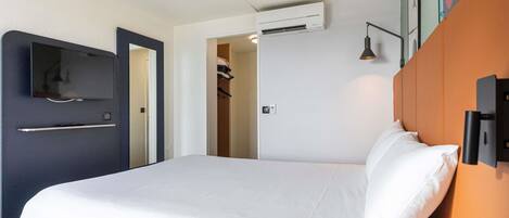 A hotel room with a bed, a TV, an air conditioning unit, a door, and a wall-mounted lamp.