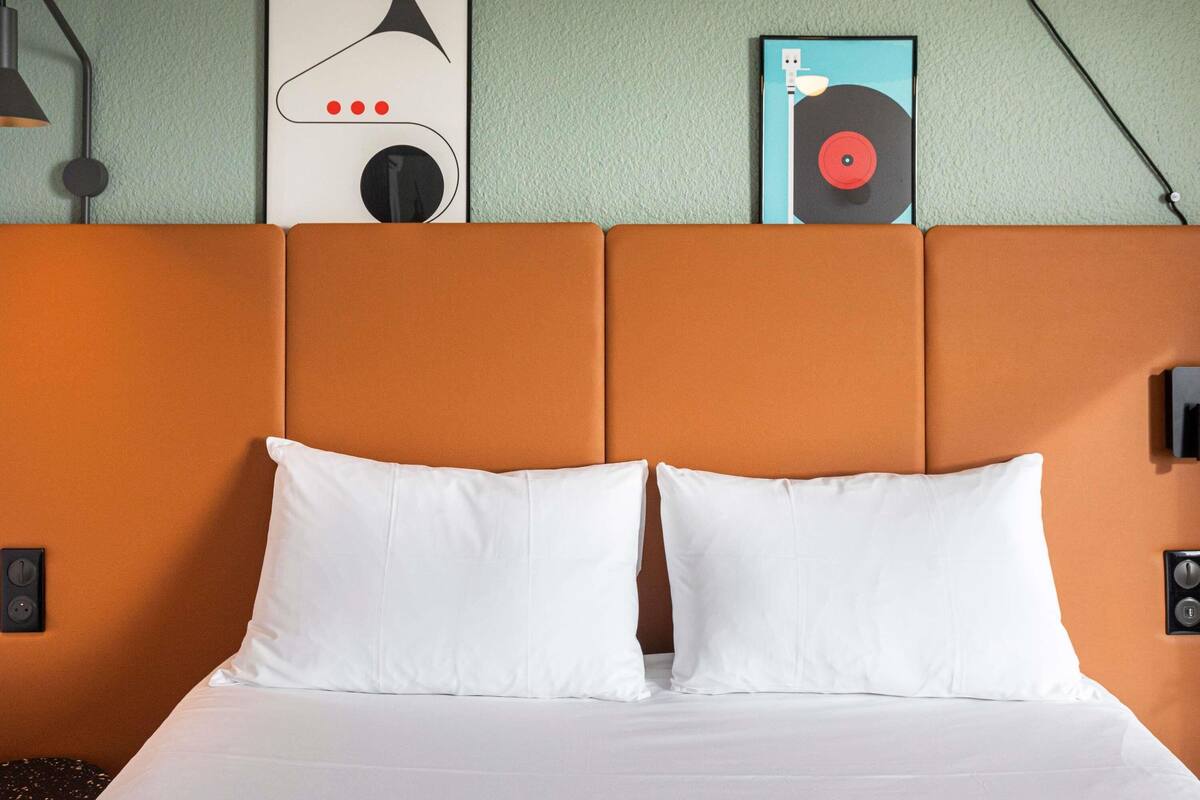 A bed with white pillows and an orange headboard, a wall-mounted light, and two framed artworks above the bed.