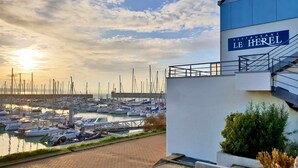 A marina with boats docked, a building with a sign reading "LE HEREL," and a clear sky with the sun setting or rising. - ibis Granville Port de plaisance (Granville)