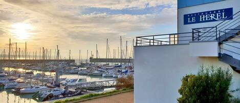A marina with boats docked, a building with a sign reading "LE HEREL," and a clear sky with the sun setting or rising.