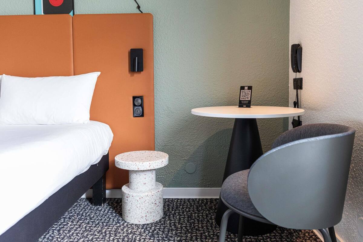 A hotel room with a bed, a small round table, a chair, a wall-mounted phone, and a QR code sign.