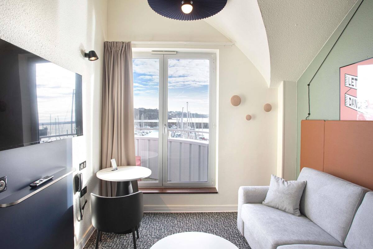 A compact hotel room with a large window, a sofa, a small table, and a TV.