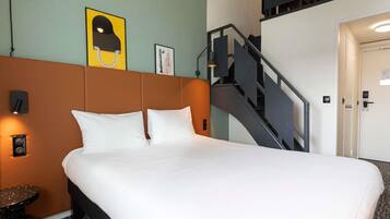 A hotel room with a large bed, a headboard, a small table, a wall-mounted lamp, a framed picture, and a staircase.