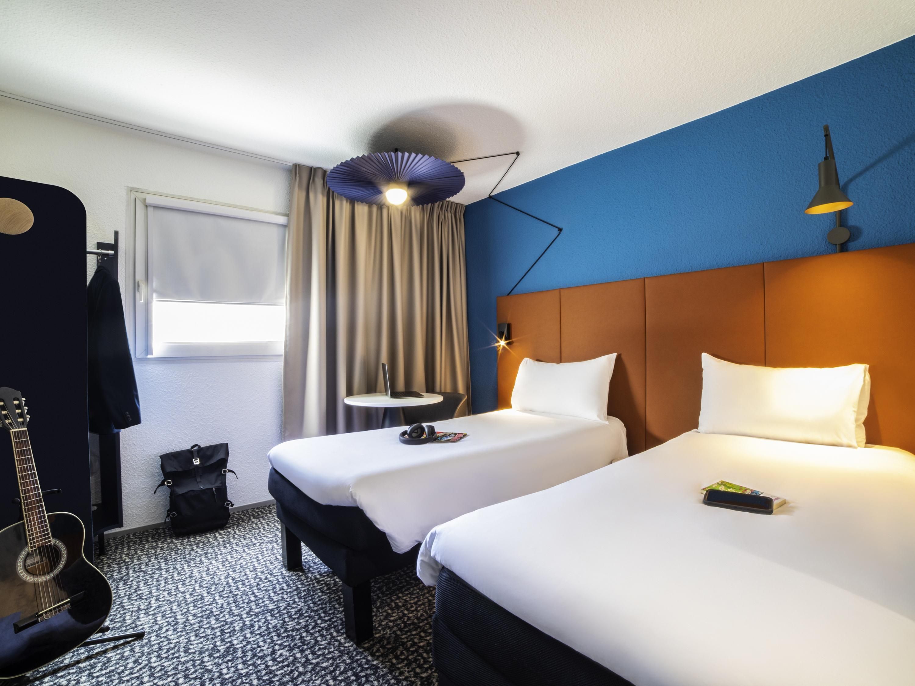Une chambre d’hôtel avec deux lits, un bureau, une guitare et une fenêtre avec des rideaux.