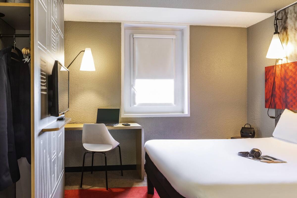 Habitación de hotel con cama, escritorio, laptop, silla, armario y ventana con persianas.