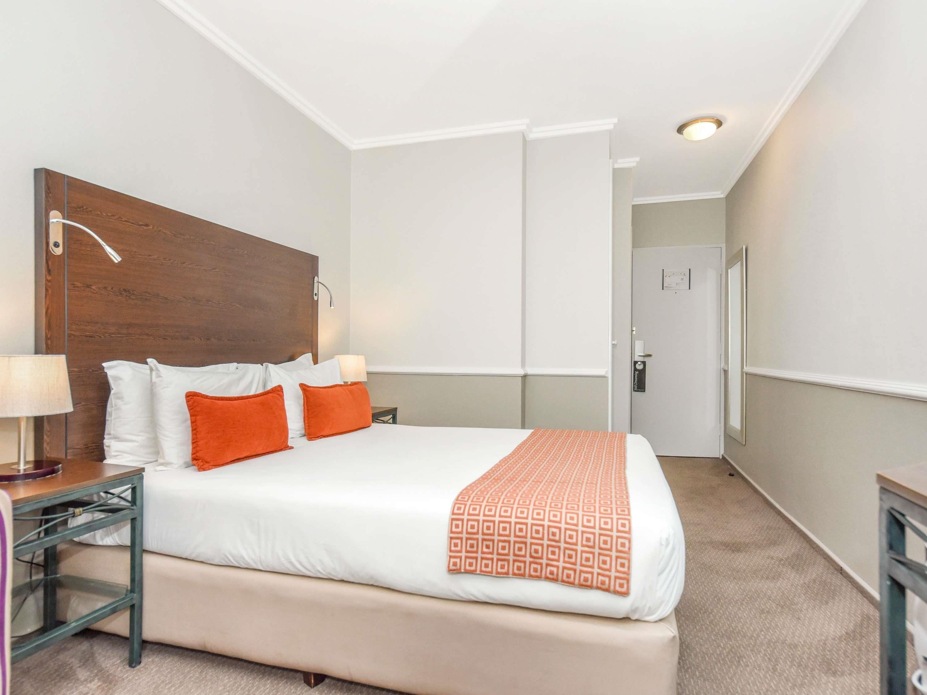 Une chambre d’hôtel avec un grand lit, du linge de lit blanc, des oreillers orange, une tête de lit en bois, une table de chevet avec une lampe et une porte avec une serrure.