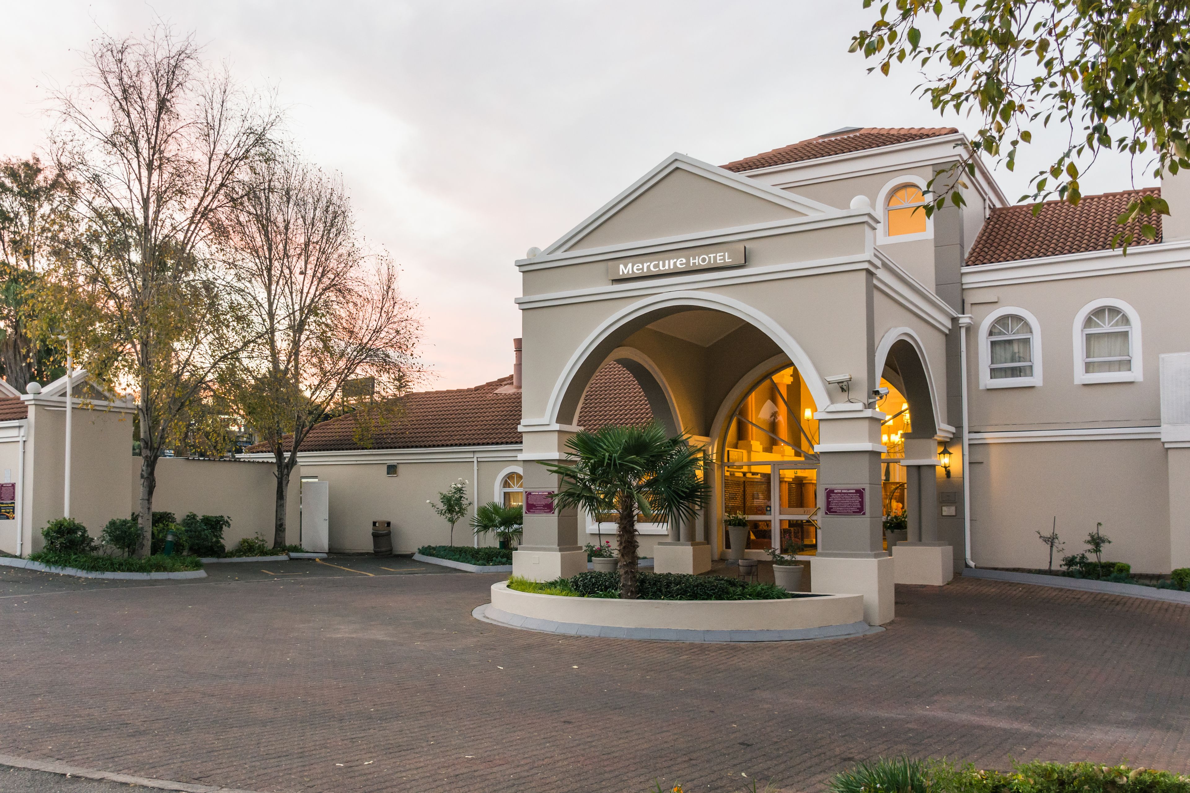 A hotel entrance with a sign reading "Mercure HOTEL," featuring a central archway, a landscaped garden, and a brick-paved driveway.
