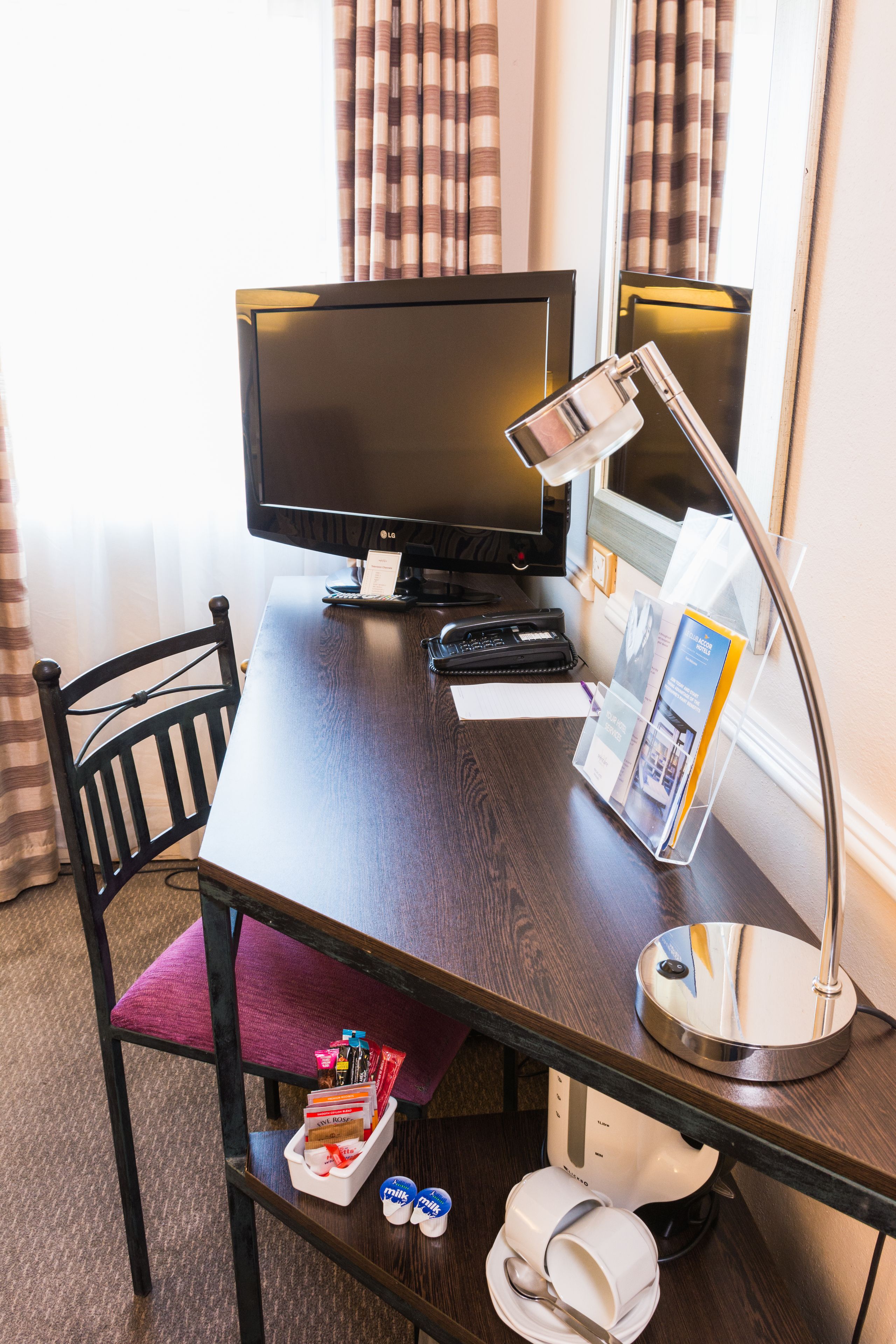 Ein Schreibtisch im Hotelzimmer mit einem Computer, einer Lampe, einem Stuhl und verschiedenen Gegenständen darauf.