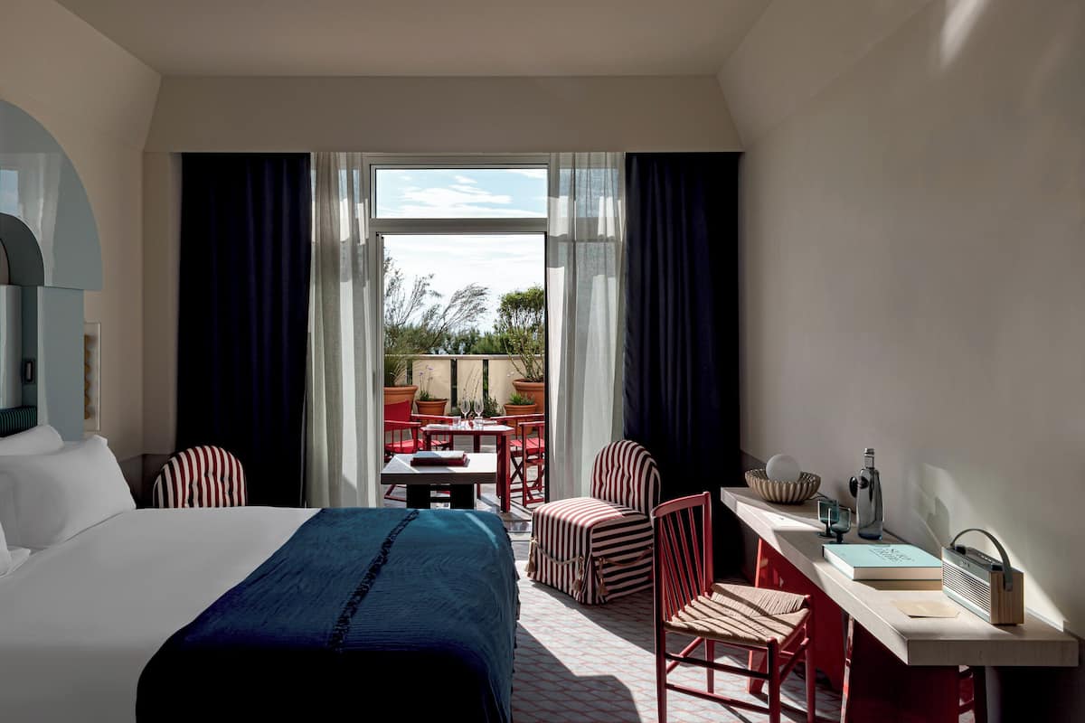 A hotel room with a bed, a desk with a radio, a chair, and a view of the outdoor patio.