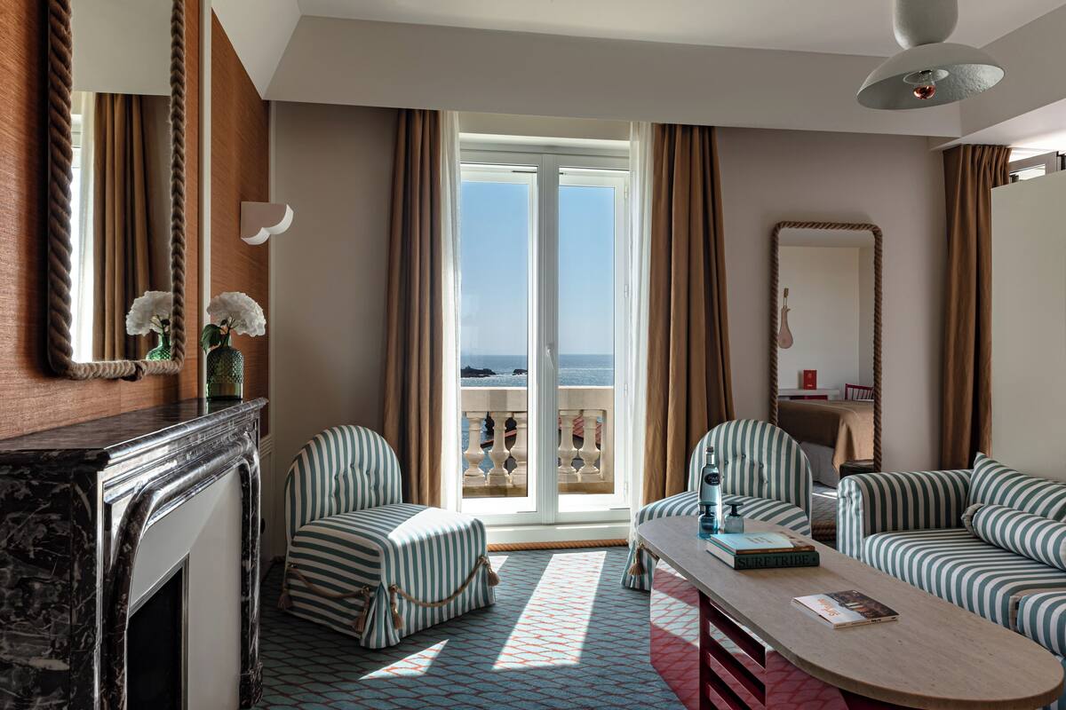 A hotel room with a balcony, striped upholstered furniture, a fireplace, and a view of the sea.