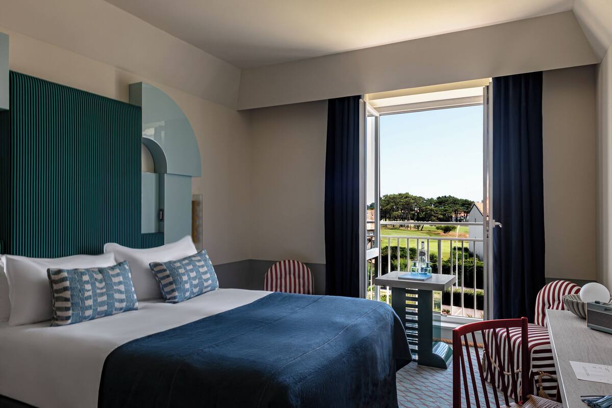 A hotel room with a bed, a small table, chairs, and a balcony with a view of the greenery.