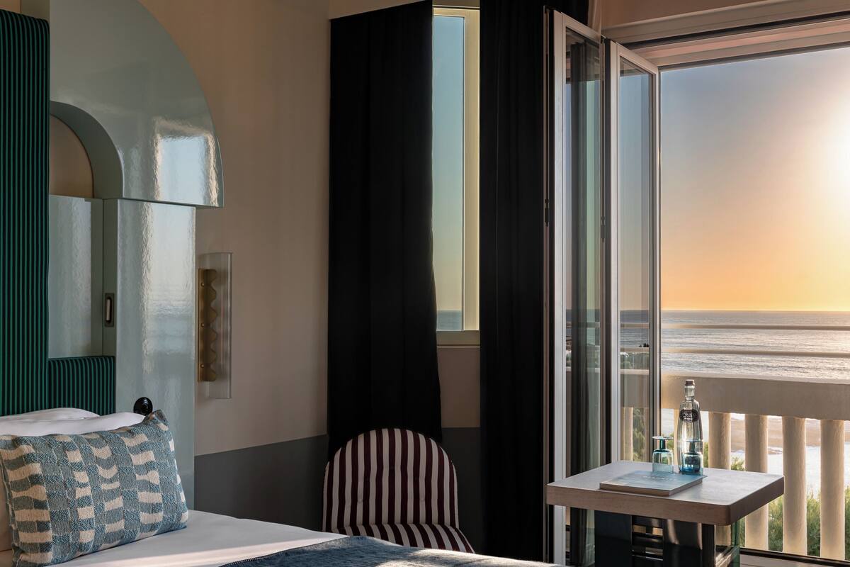 A hotel room with a bed, a chair, a table with a bottle, and a view of the ocean.