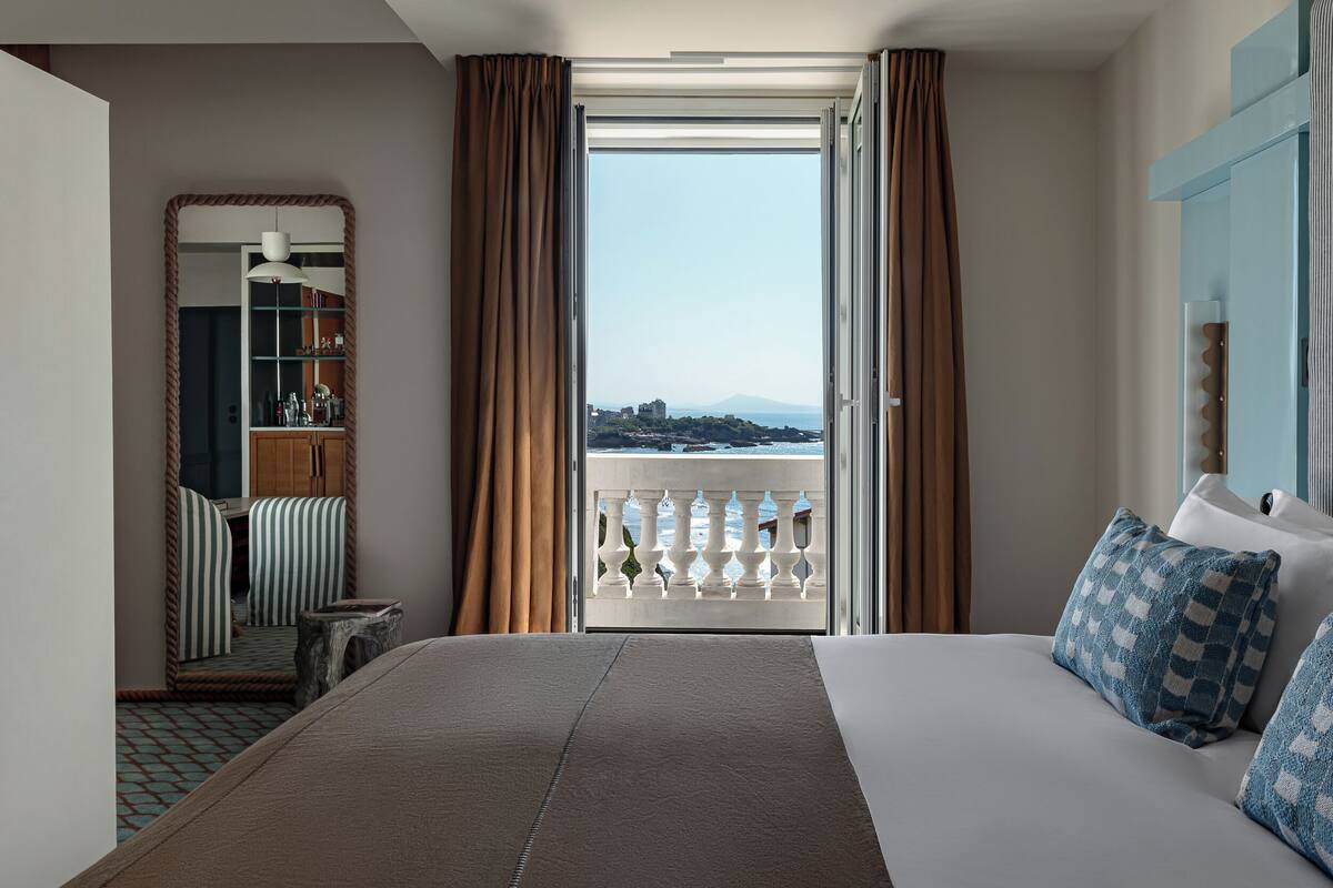 A hotel room with a large bed, a view of the city through an open window, a balcony with a railing, and a seating area with a chair and a small table.