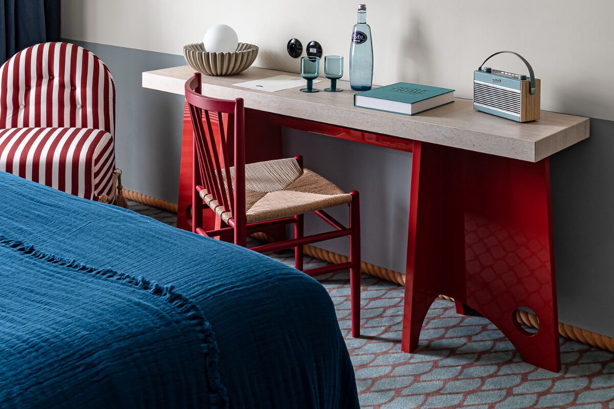 Een hotelkamer met een bed, een stoel met rode en witte strepen, een rode bureau met een radio, en een blauw boek.