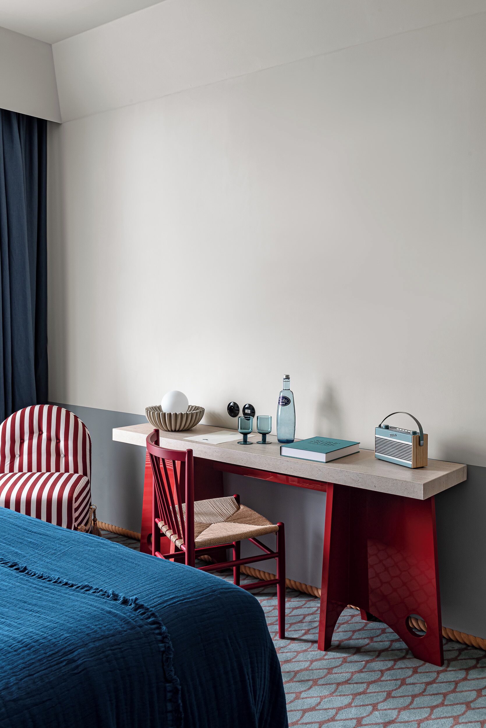 A hotel room with a bed, a desk with a chair, a radio, a book, a bottle, and a lamp.