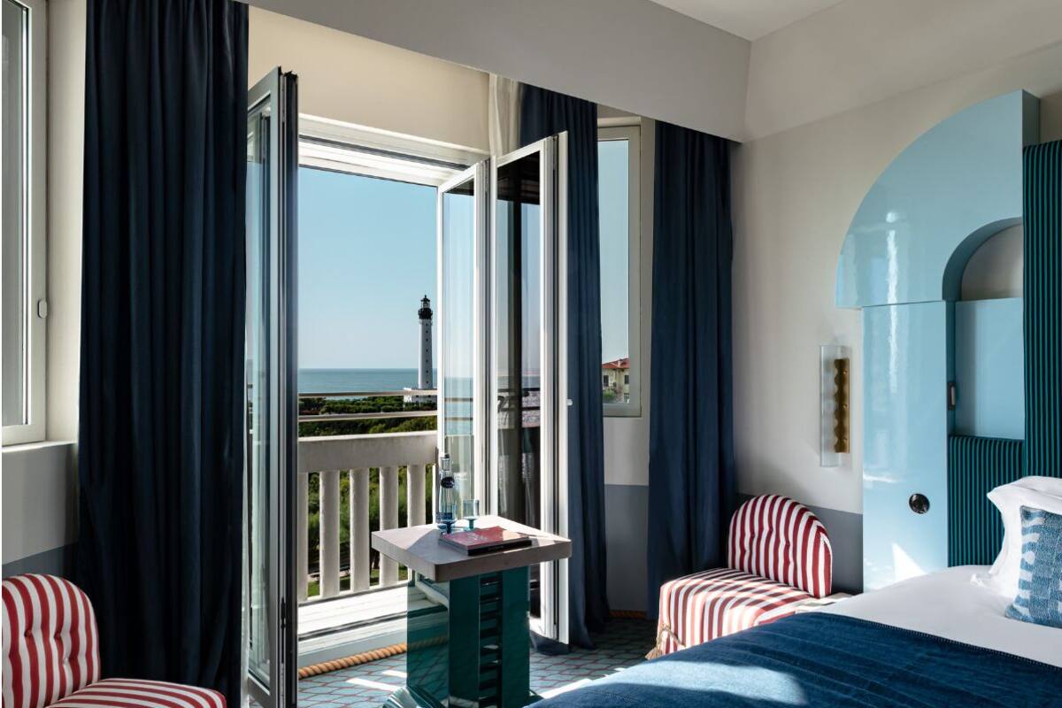 A hotel room with a balcony, a bed, a small table, and striped chairs.