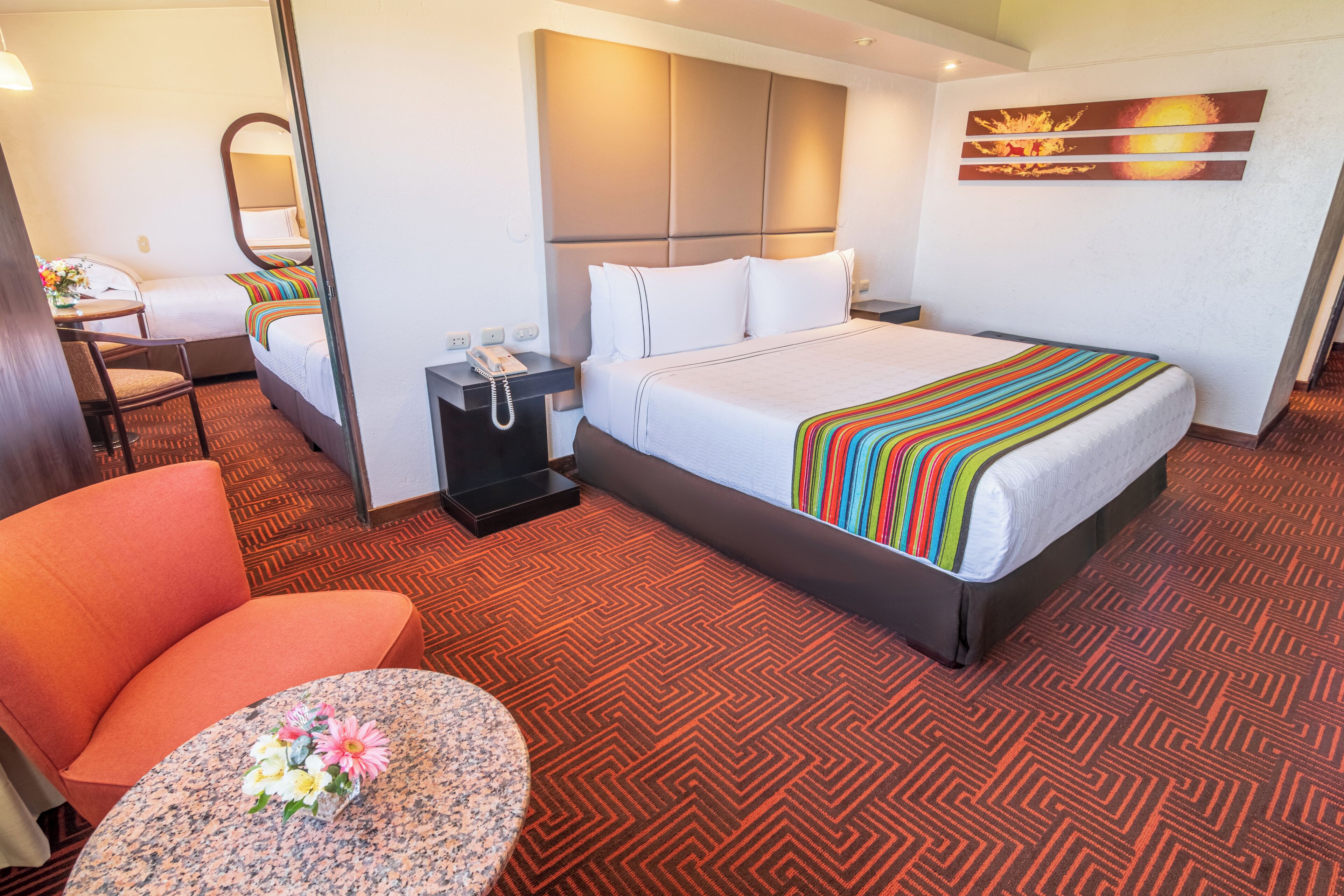 Ein Hotelzimmer mit einem Bett, einem Schreibtisch, einem Sessel, einem kleinen Tisch mit Blumen und einem an der Wand hängenden Kunstwerk.