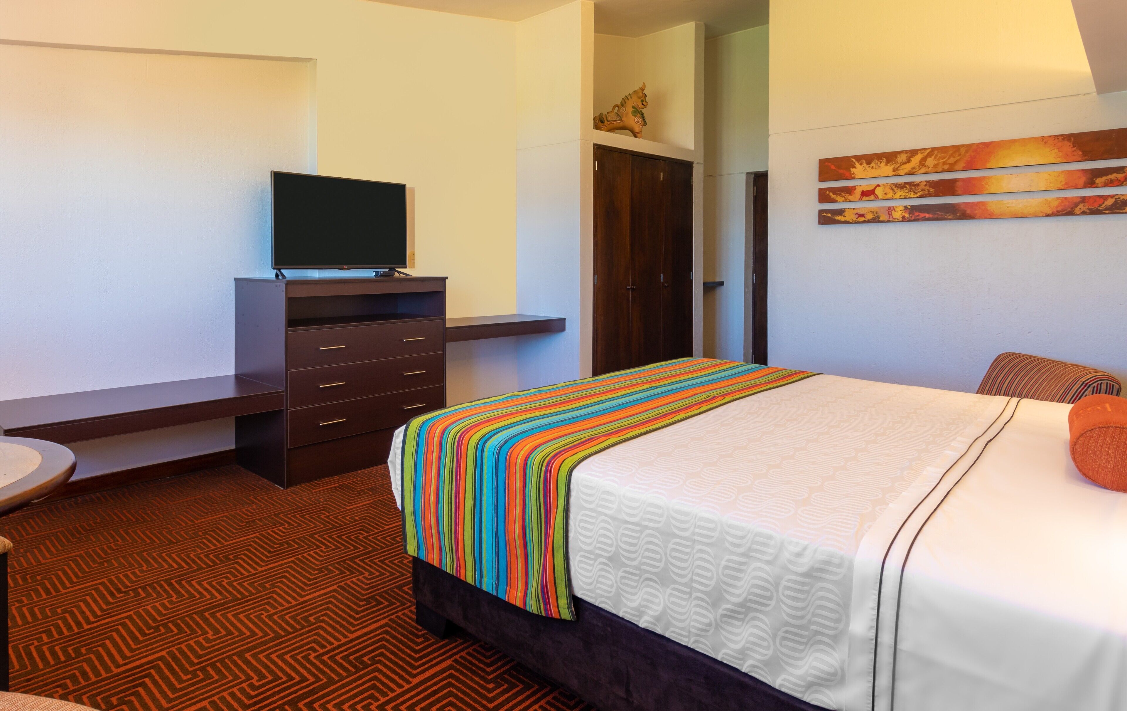 A hotel room with a bed, a TV on a dresser, a wardrobe, and a painting on the wall.