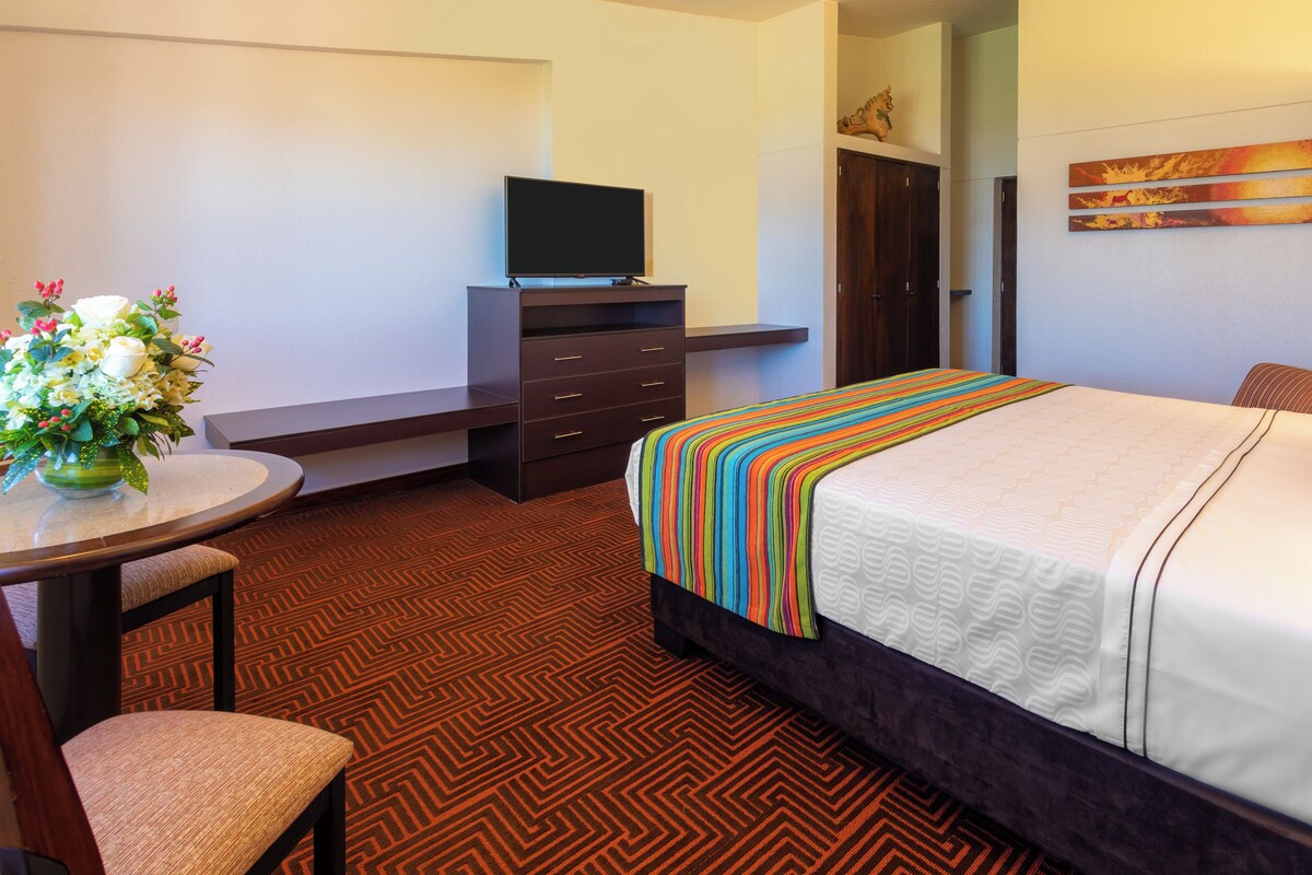A hotel room with a bed, a desk with a chair, a television, and a painting on the wall.