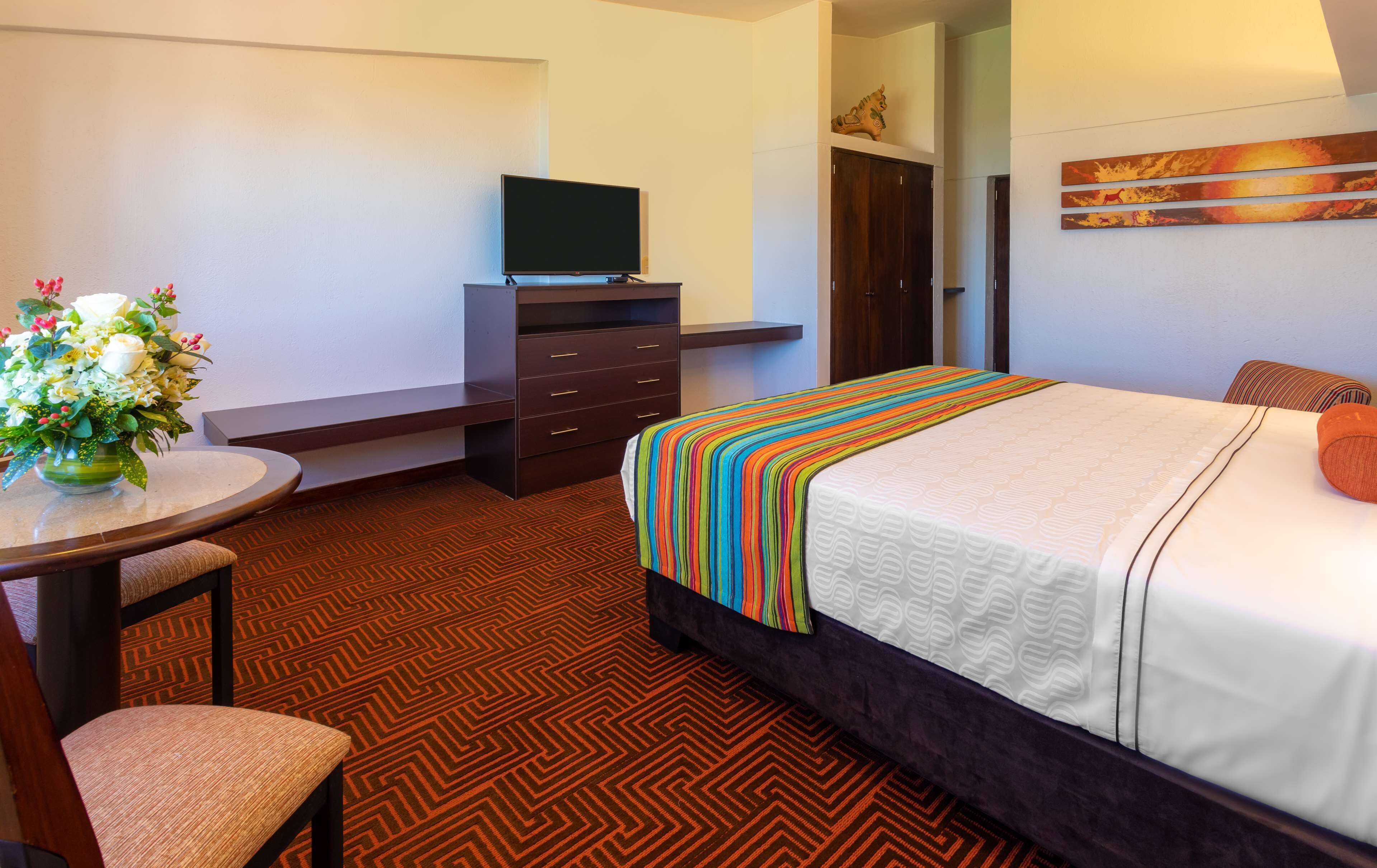 Une chambre d’hôtel comprenant un lit, un bureau avec une chaise, une télévision, une commode et un tableau au mur.